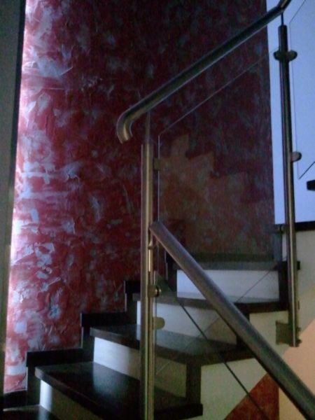 Schody z ciemnego drewna z nowoczesną balustradą ze stali nierdzewnej i szkła, na tle ściany z dekoracyjnym, czerwono-białym tynkiem.