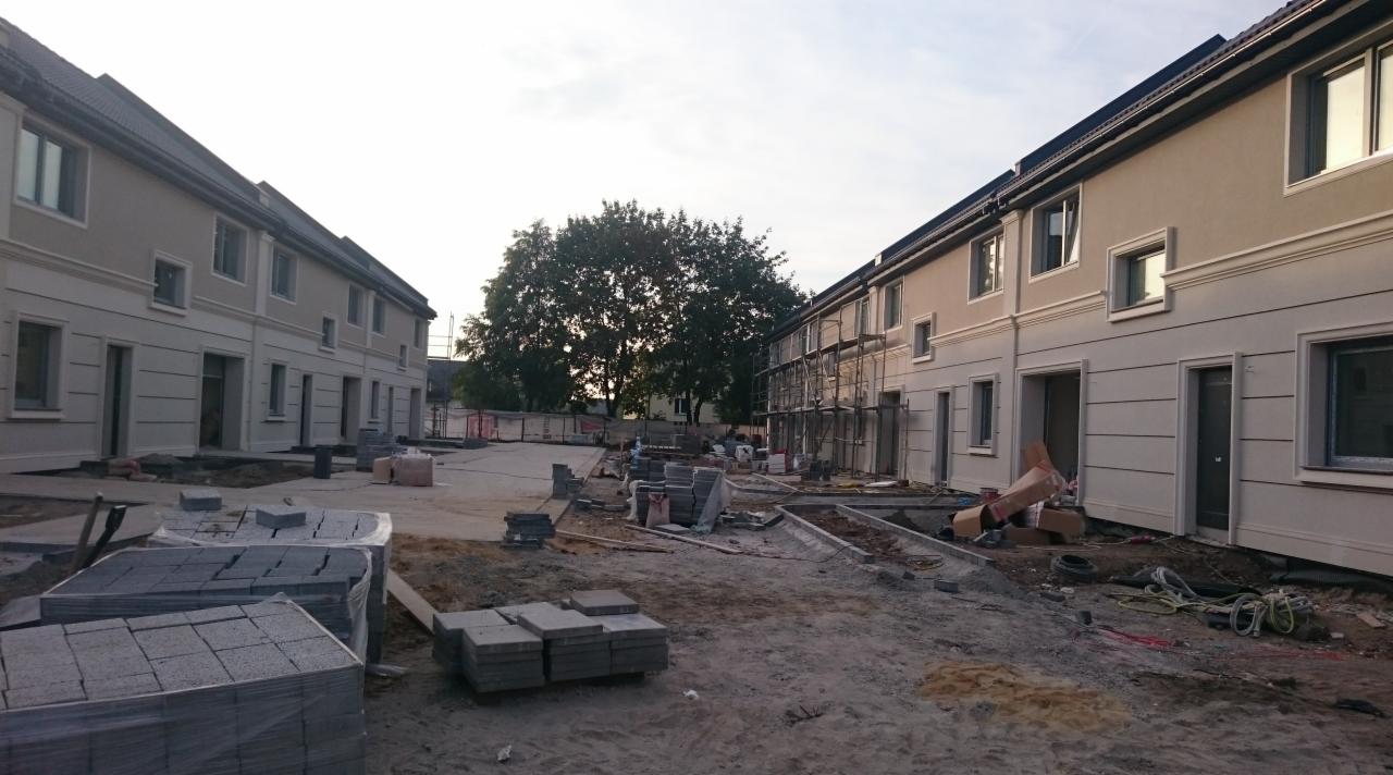 Domy szeregowe w trakcie prac wykończeniowych elewacji, widoczne rusztowania, przygotowane materiały budowlane i teren budowy.