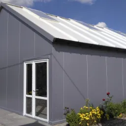 Gotowy budynek z płyt warstwowych w szarym kolorze z białym dachem i oknami dachowymi, otoczony zielenią i kwiatami, z fragmentem samochodu po lewej stronie.