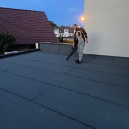 ART-DACH Dawid Mikołajczak - Pracownik używa dmuchawy do czyszczenia płaskiego dachu pokrytego papą w Oleśnicy, w tle widoczny budynek z czerwoną dachówką.