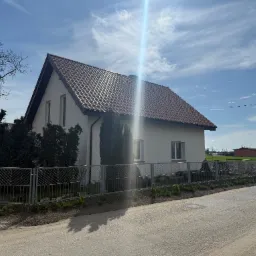 Dom jednorodzinny z brązowym dachem z dachówki ceramicznej, ogrodzony metalowym płotem, w słoneczny dzień. Widoczne drzewa i fragment drogi.