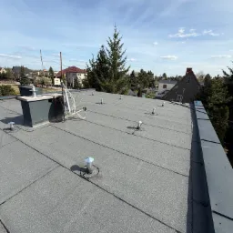 Szary, płaski dach pokryty papą termozgrzewalną z kominem i odpowietrznikami na tle zabudowy jednorodzinnej i błękitnego nieba.