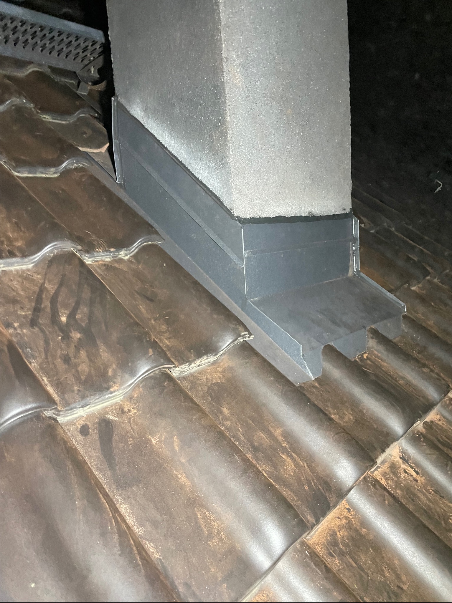 Szare, metalowe wykończenie komina na brązowym dachu z dachówki ceramicznej, widoczne elementy wentylacyjne.