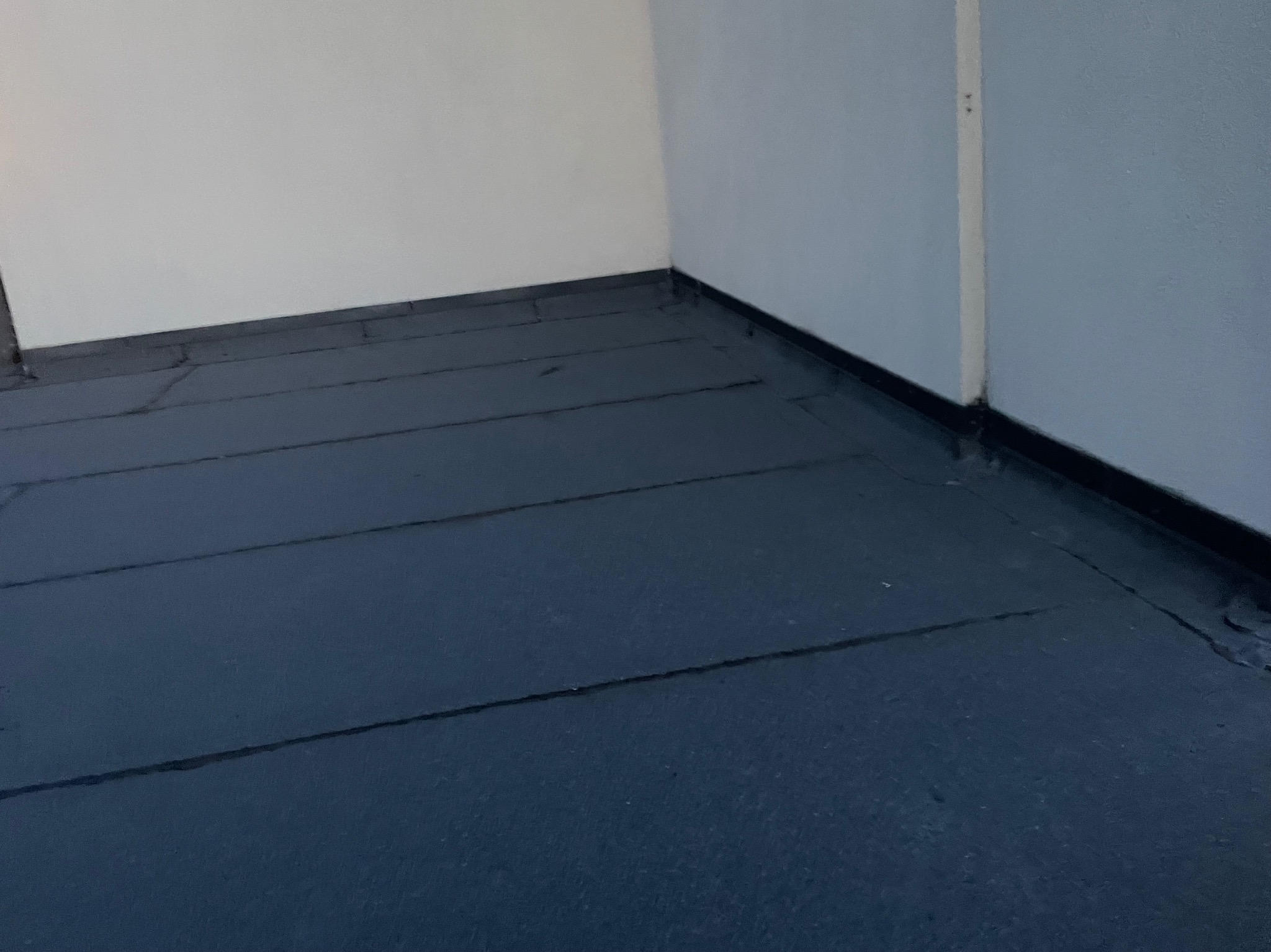 Fragment dachu płaskiego pokrytego papą termozgrzewalną, widoczne łączenia papy przy ścianie budynku, w tle biała i szara ściana.