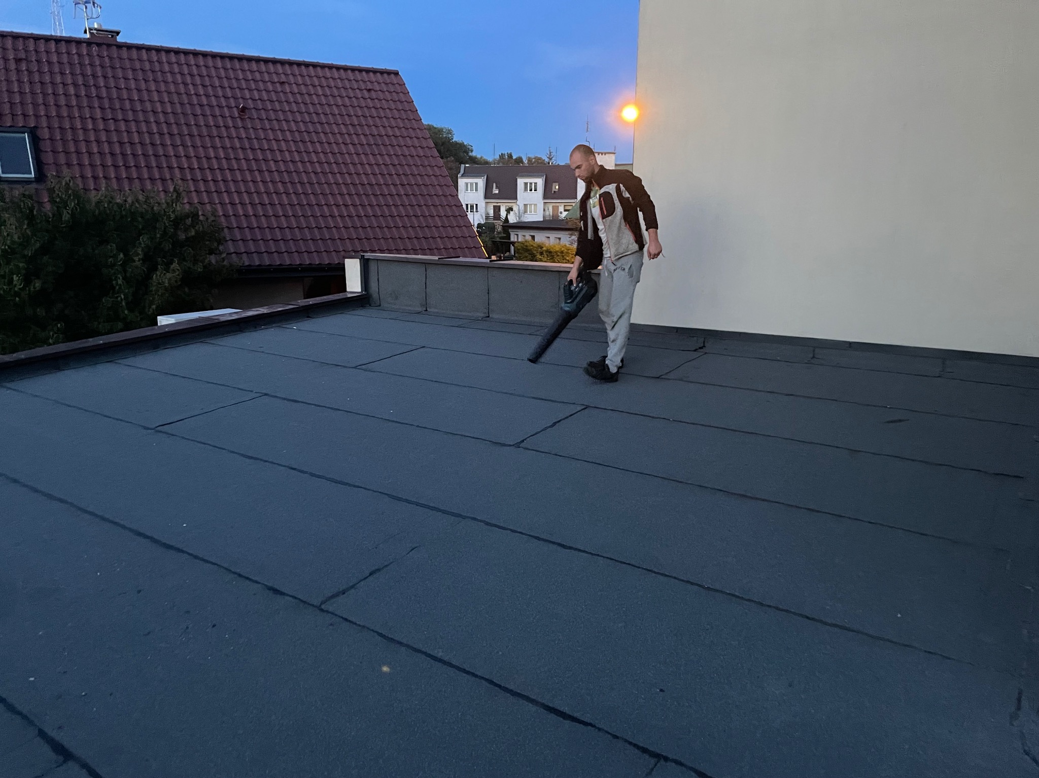 Pracownik używa dmuchawy do czyszczenia płaskiego dachu pokrytego papą w Oleśnicy, w tle widoczny budynek z czerwoną dachówką.