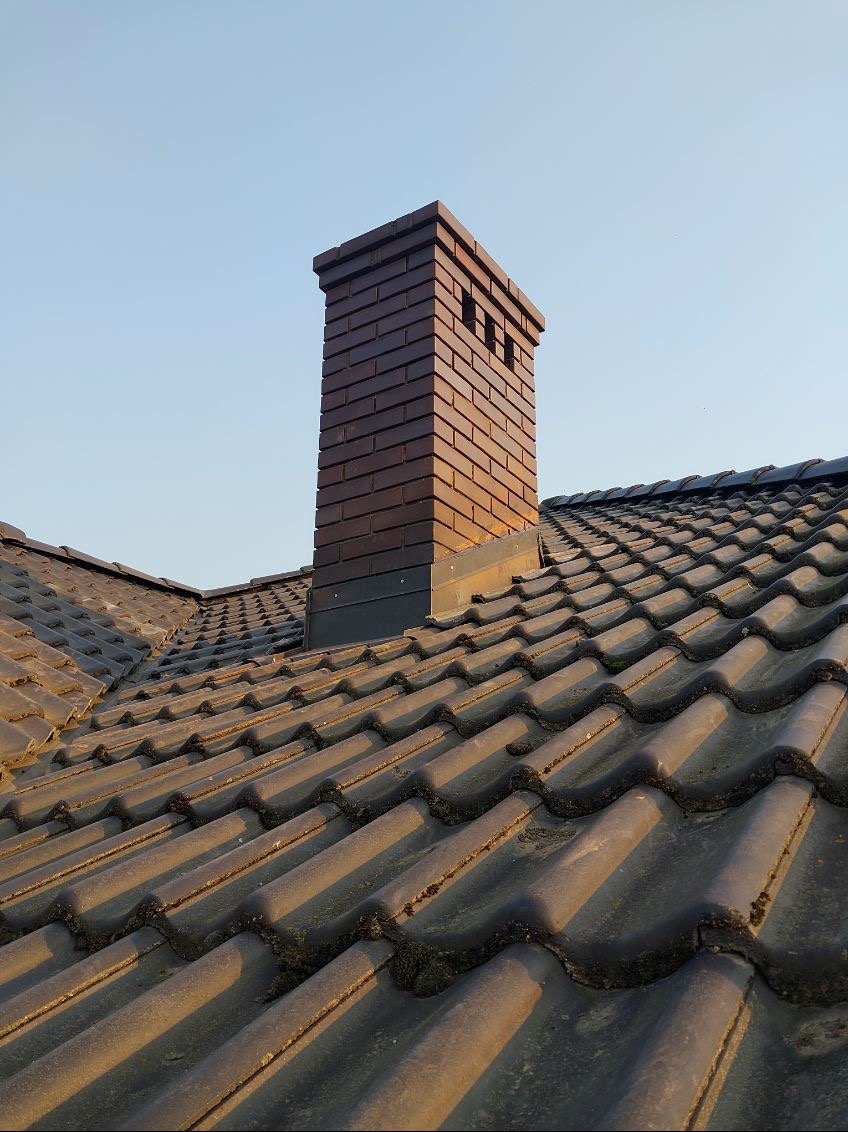 Ciemnobrązowy komin z cegły na dachu pokrytym dachówką ceramiczną w Kaliszu, widoczne ślady mchu na dachówce.