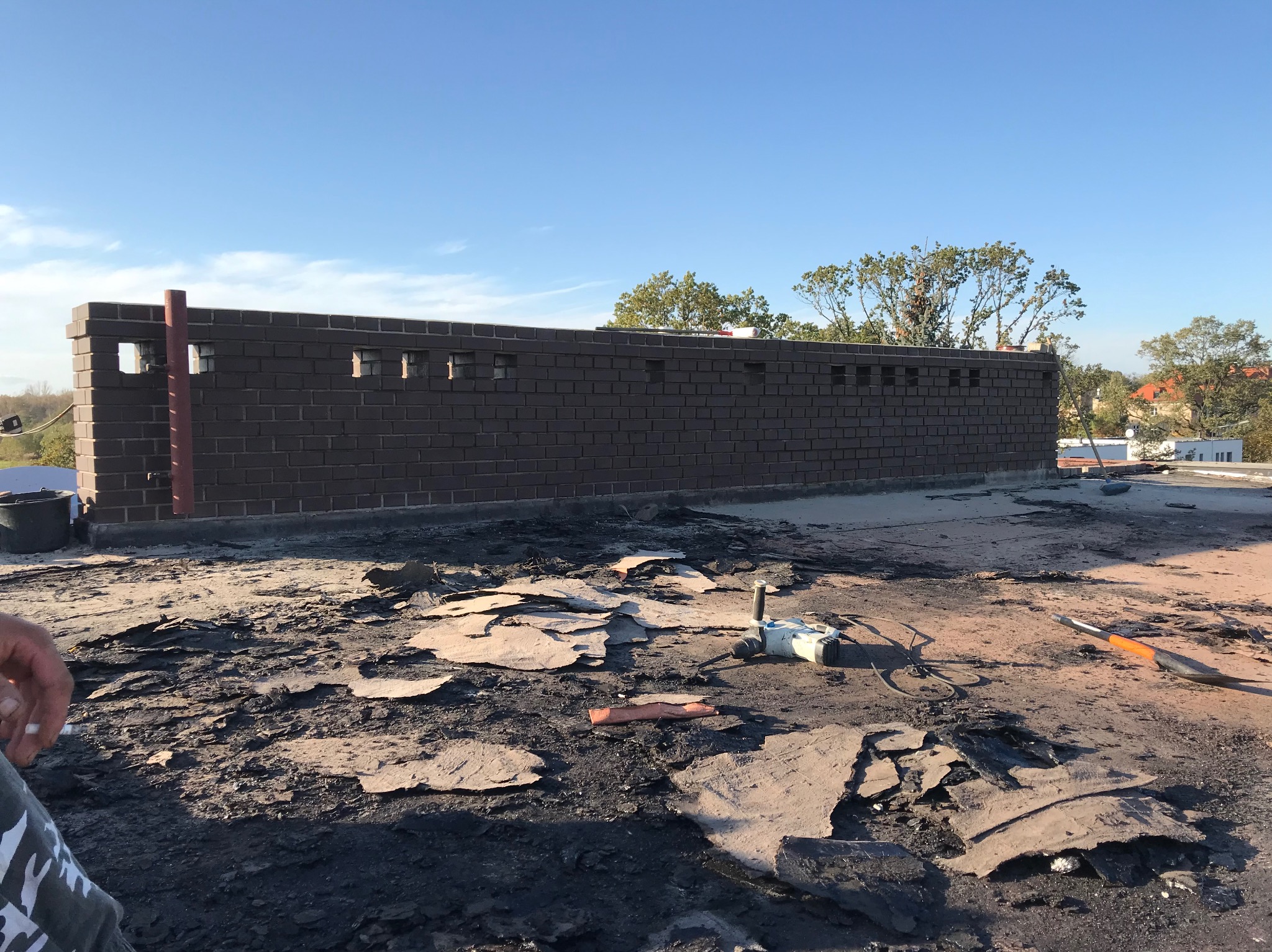 Demontaż starego pokrycia dachowego na płaskim dachu, widoczny fragment muru attyki z cegły klinkierowej i narzędzia pracy. Lokalizacja Wrocław Zakopiańska.