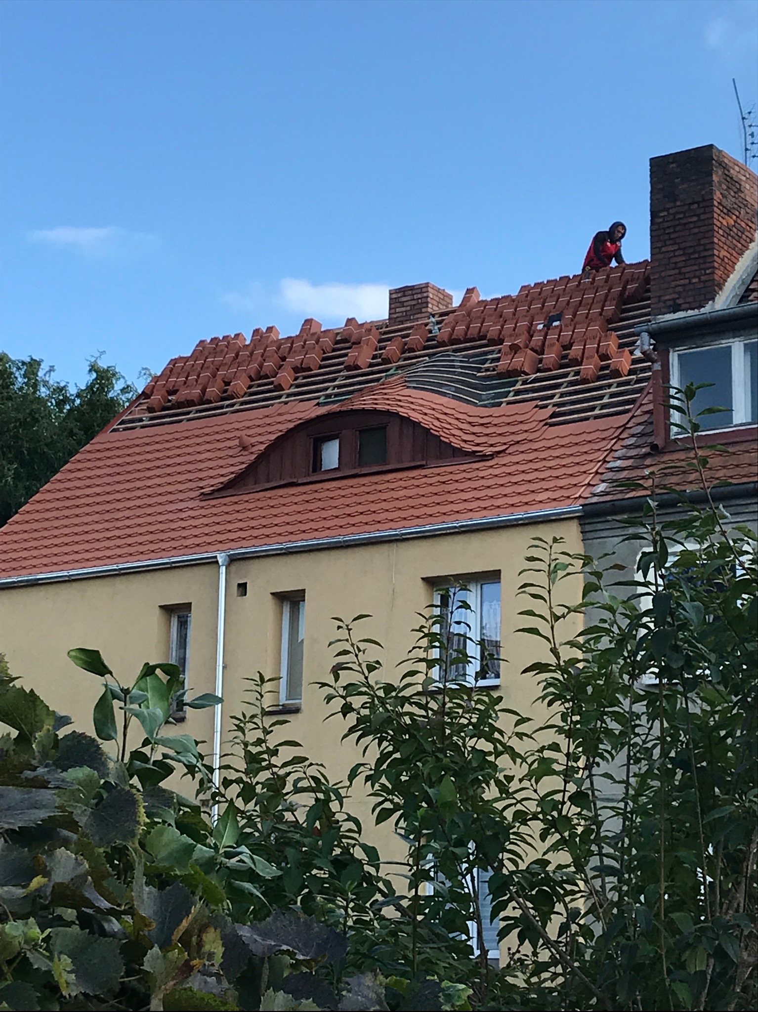 Częściowo zdjęty dach z czerwonej dachówki na żółtym budynku mieszkalnym we Wrocławiu, dekarz w czerwonej koszulce układa nową dachówkę.