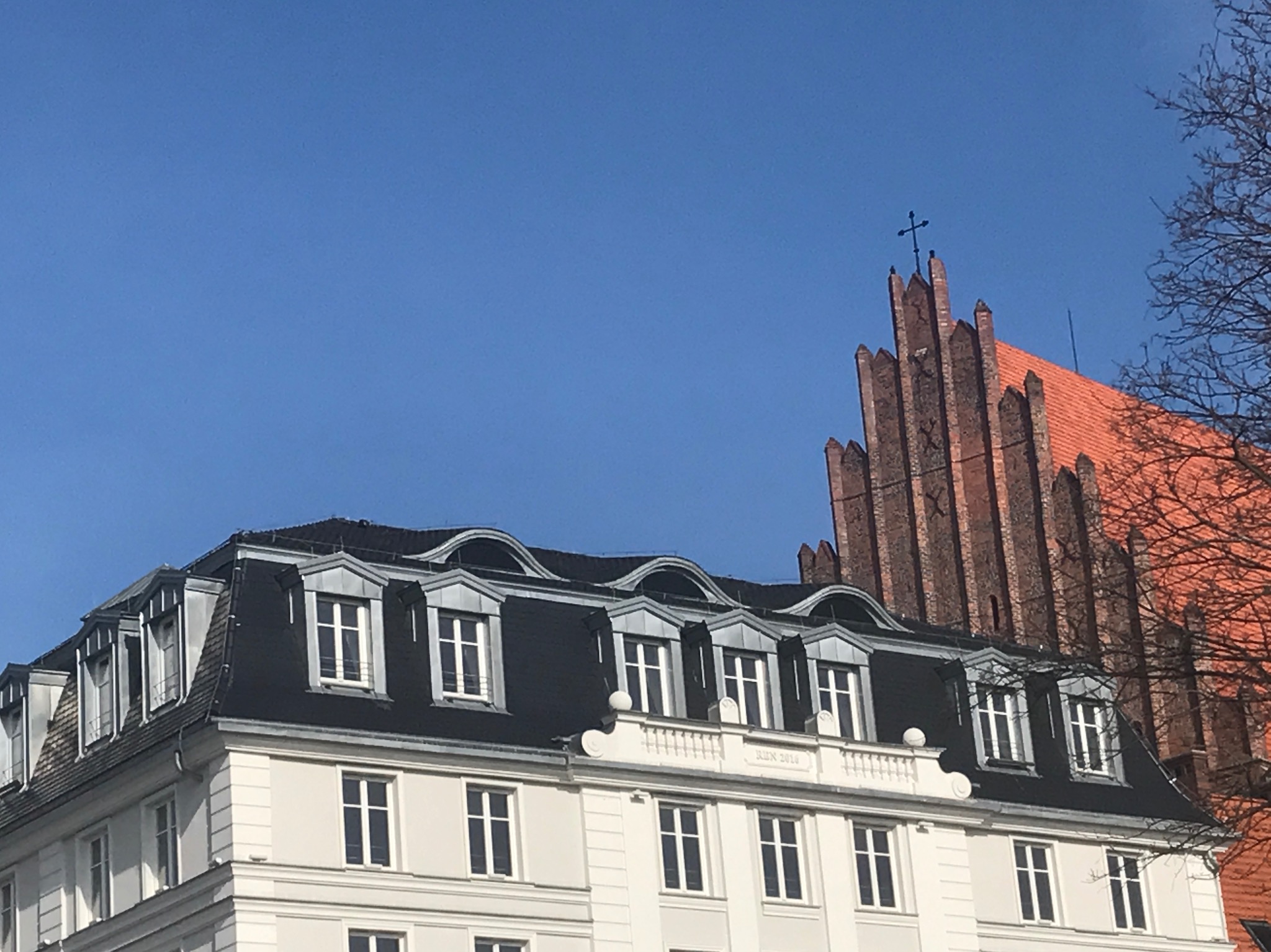 Fragment kamienicy we Wrocławiu, pl. Wolności, z czarnym dachem i lukarnami, na tle ceglanej wieży kościelnej z krzyżem.