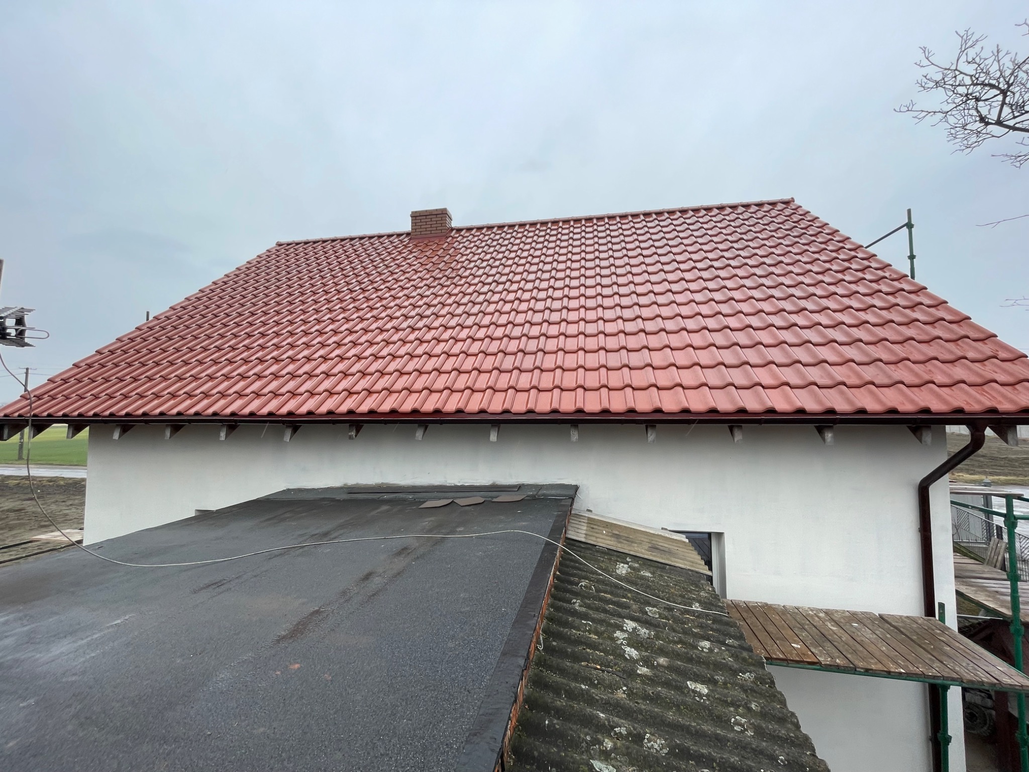 Widok z góry na dom z nowym, czerwonym dachem z dachówki ceramicznej, z kominem z cegły, kontrastujący z fragmentami starego, bitumicznego i eternitowego pokrycia dachu na pierwszym planie.