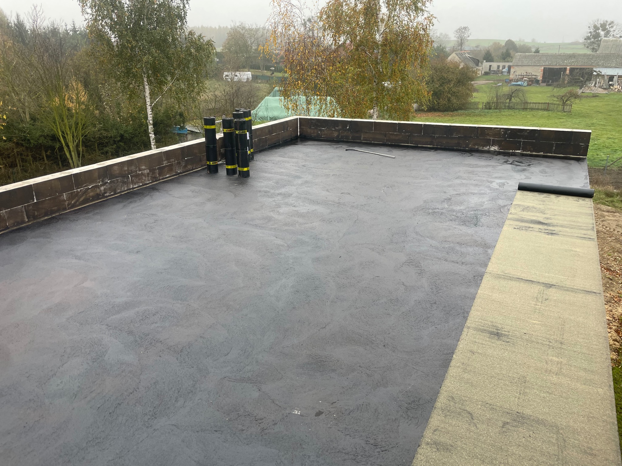 Płaski dach pokryty świeżą, czarną hydroizolacją bitumiczną, z widocznymi rolkami papy i narzędziem, na tle jesiennego krajobrazu wiejskiego.