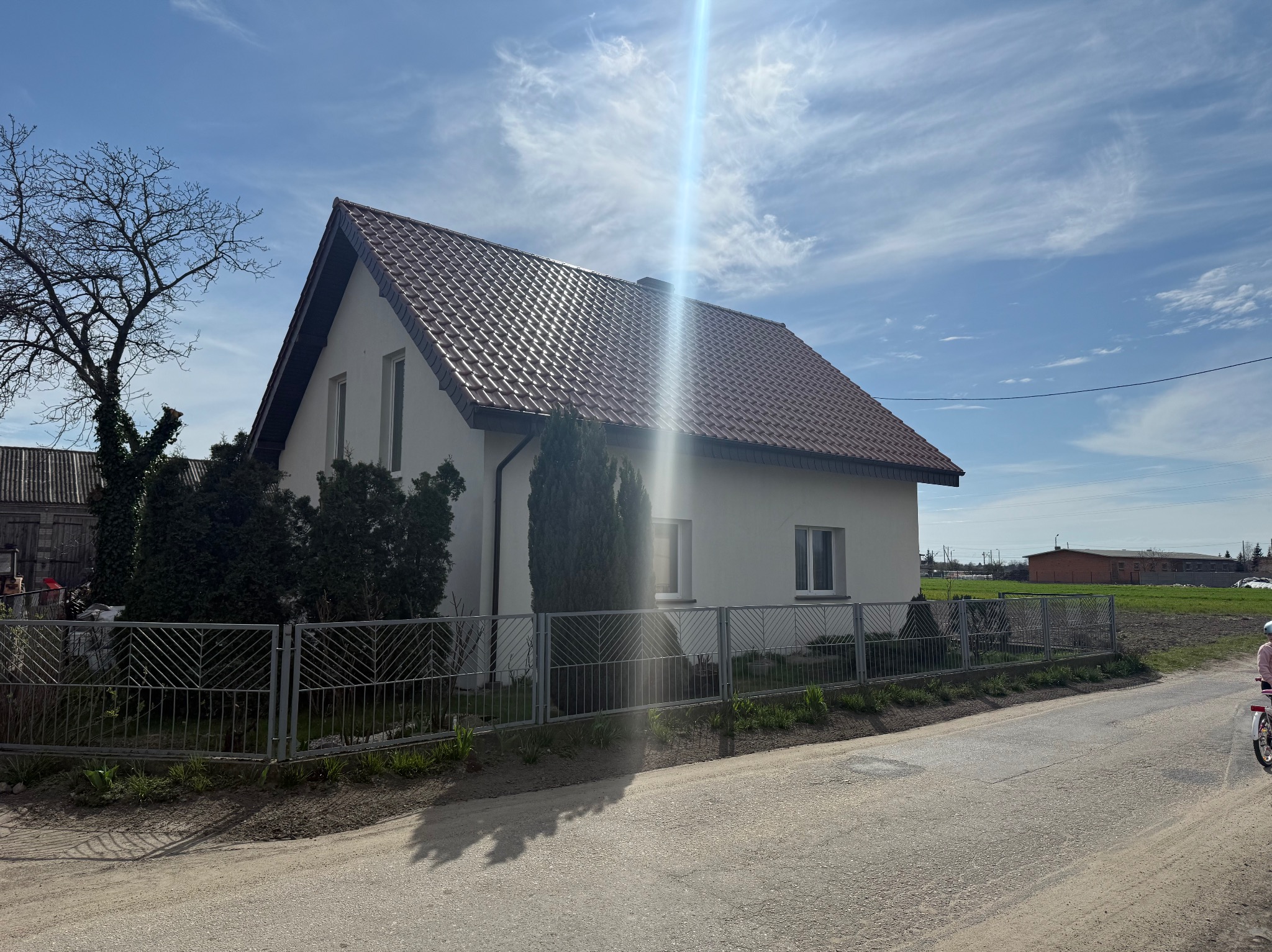 Dom jednorodzinny z brązowym dachem z dachówki ceramicznej, ogrodzony metalowym płotem, w słoneczny dzień. Widoczne drzewa i fragment drogi.