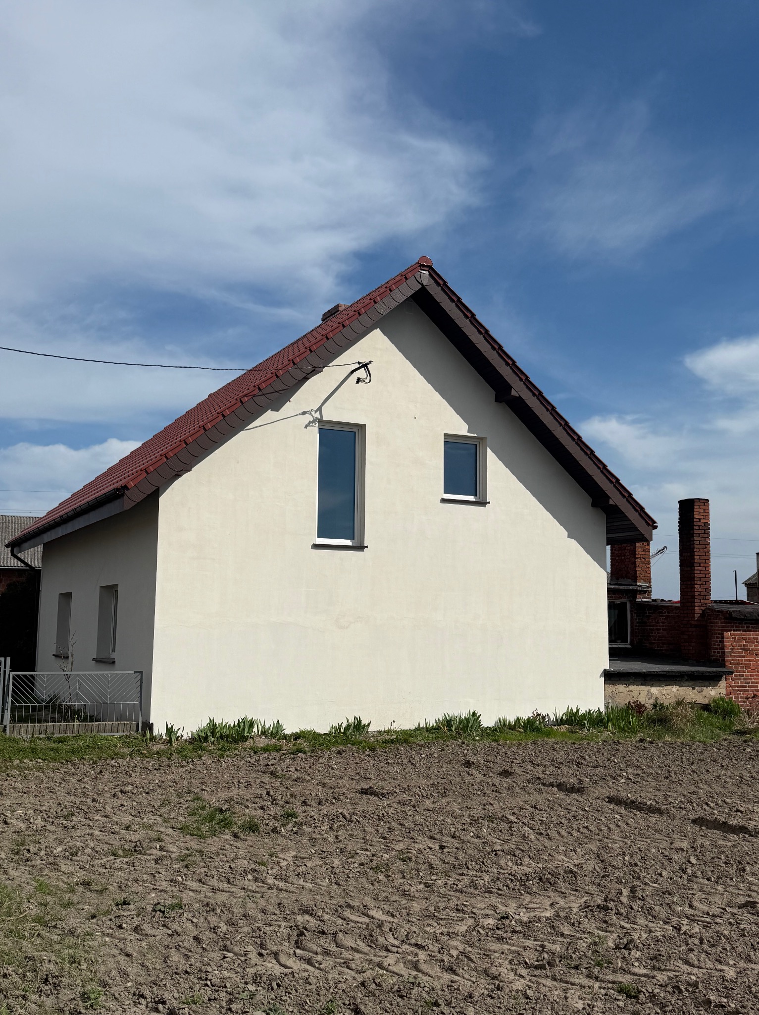 Biały dom jednorodzinny z czerwoną dachówką na tle błękitnego nieba z chmurami. Widoczne okna o różnej wysokości i przygotowane podłoże pod trawnik.