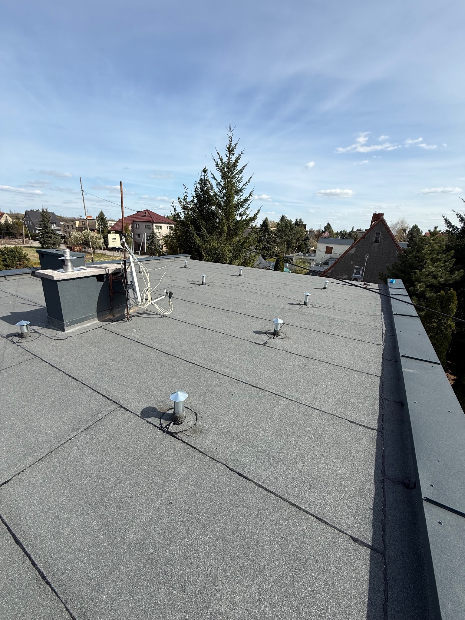 Szary, płaski dach pokryty papą termozgrzewalną z kominem i odpowietrznikami na tle zabudowy jednorodzinnej i błękitnego nieba.