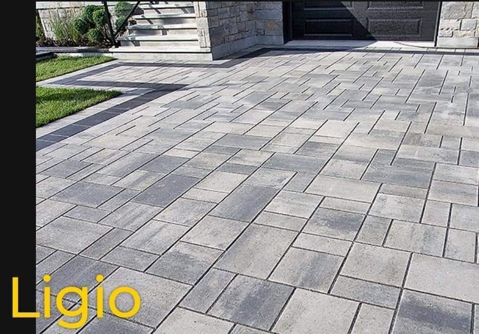 Szara nawierzchnia z prostokątnych elementów ułożonych w nieregularny wzór, obrzeże z ciemniejszych elementów, schody wejściowe do domu w tle.