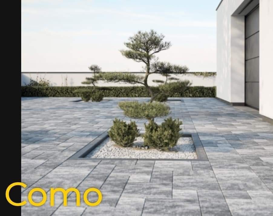 Nowoczesna nawierzchnia z szarej kostki, z wkomponowanymi rabatami z białym żwirem i formowanymi drzewkami, na tle białego muru i budynku.