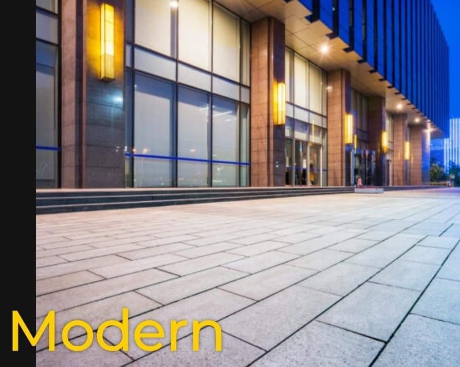 Nowoczesna nawierzchnia z jasnoszarej kostki betonowej, ułożonej w prostokątny wzór przed wejściem do budynku biurowego z dużymi oknami i oświetleniem zewnętrznym.