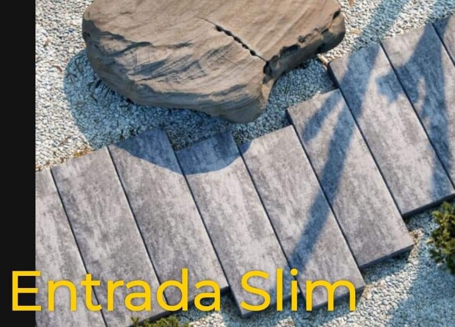 Ścieżka z szarych płyt ułożonych w ogrodzie z jasnym żwirem, obok duży, nieregularny kamień. Widoczny napis 'Entrada Slim'.
