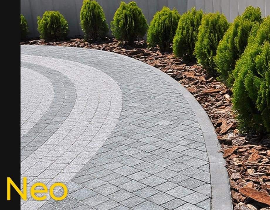 Ułożona z szarej kostki nawierzchnia w kształcie łuku, obrzeże z szarego betonu, tuje i kora drzewna w tle, widoczne logo w rogu.