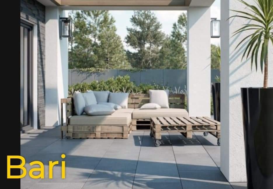 Zadaszony taras z meblami ogrodowymi wykonanymi z palet, szarymi płytami na podłodze i roślinami w tle.