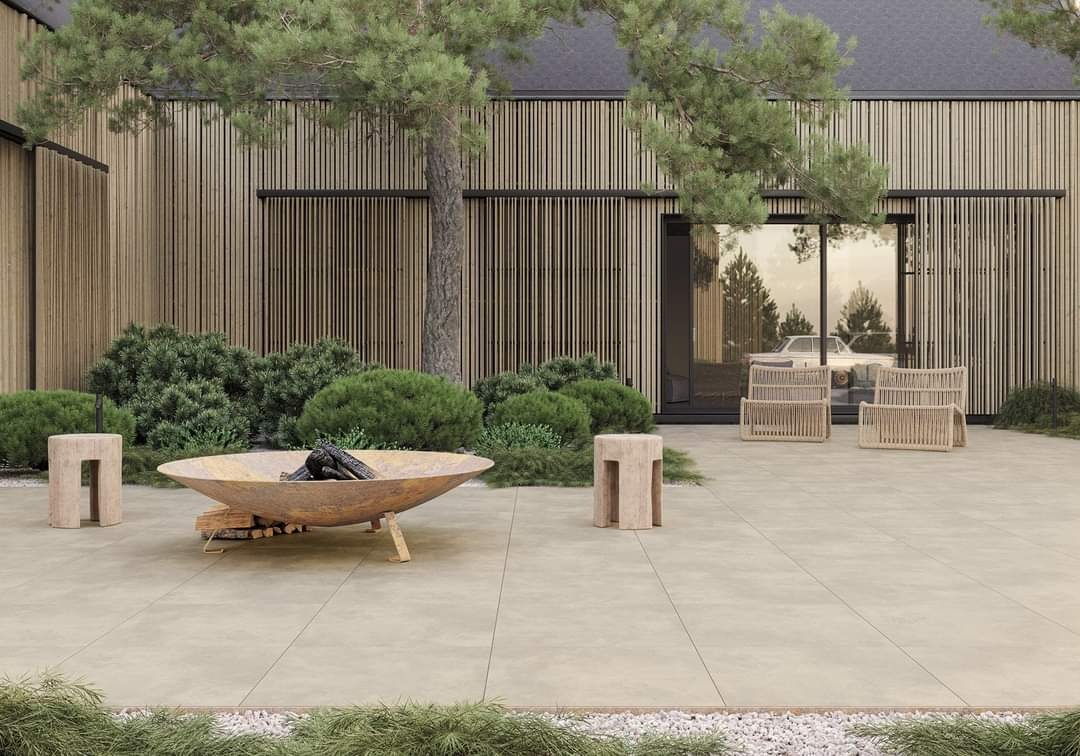 Nowoczesny taras z jasnych płyt, palenisko ogrodowe z palącym się drewnem, drewniane siedziska i ściana z pionowych desek w tle.