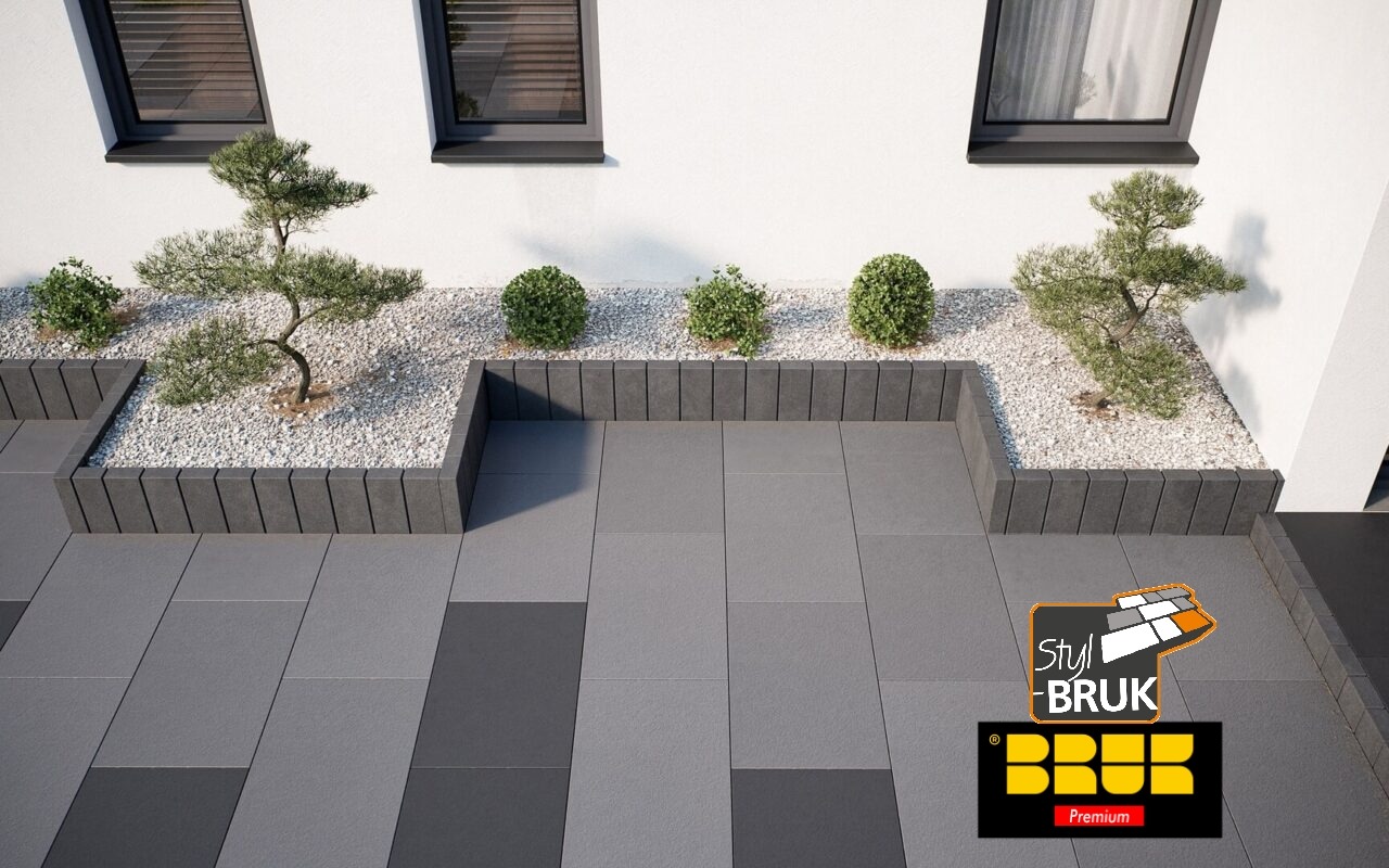 Nowoczesna nawierzchnia z szarej i grafitowej kostki, obrzeża z elementów betonowych, rabata z białym kamieniem i ozdobnymi krzewami przy białym budynku z czarnymi oknami.