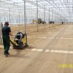 Prace ziemne w szklarni w Gdańsku: pracownik w zielonym kombinezonie obsługuje zagęszczarkę płytową, w tle widoczny ciągnik i koparka w dużej, metalowej konstrukcji.