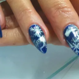 Dłoń z pomalowanymi na ciemny niebieski kolor paznokciami, dwa z nich zdobione srebrnymi płatkami śniegu i brokatem, ułożona na szklanym blacie.