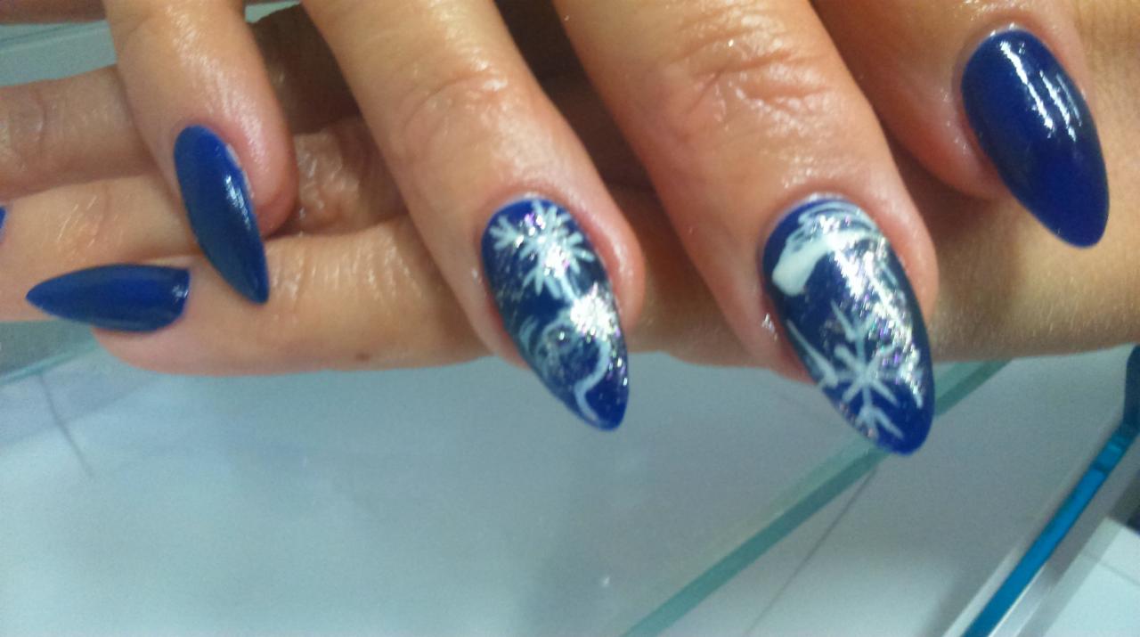 Dłoń z pomalowanymi na ciemny niebieski kolor paznokciami, dwa z nich zdobione srebrnymi płatkami śniegu i brokatem, ułożona na szklanym blacie.