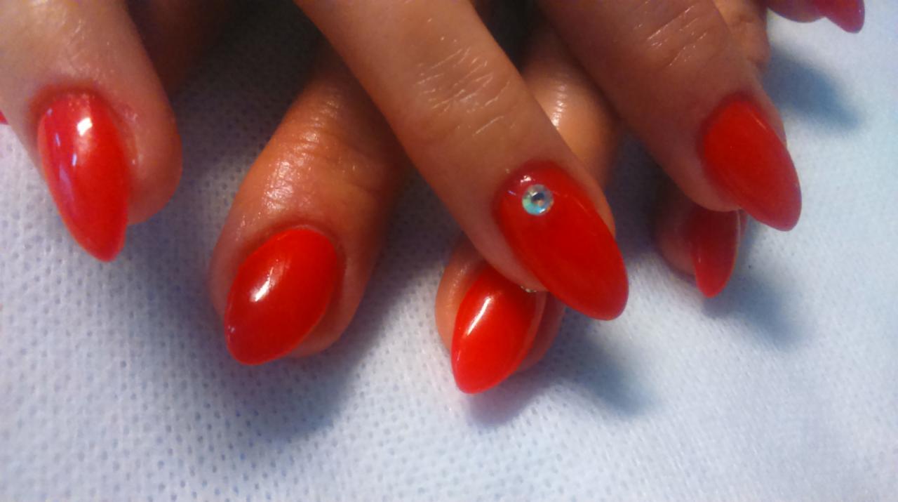 Zbliżenie na dłoń z czerwonymi, migdałowymi paznokciami; jeden paznokieć ozdobiony małym, błyszczącym dżetem.