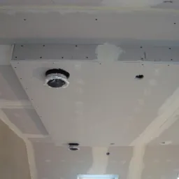 Podwieszany sufit z płyt gipsowych, widoczne łączenia i otwory na oświetlenie, w trakcie szpachlowania. Ujęcie z dołu, z widocznym fragmentem okna.