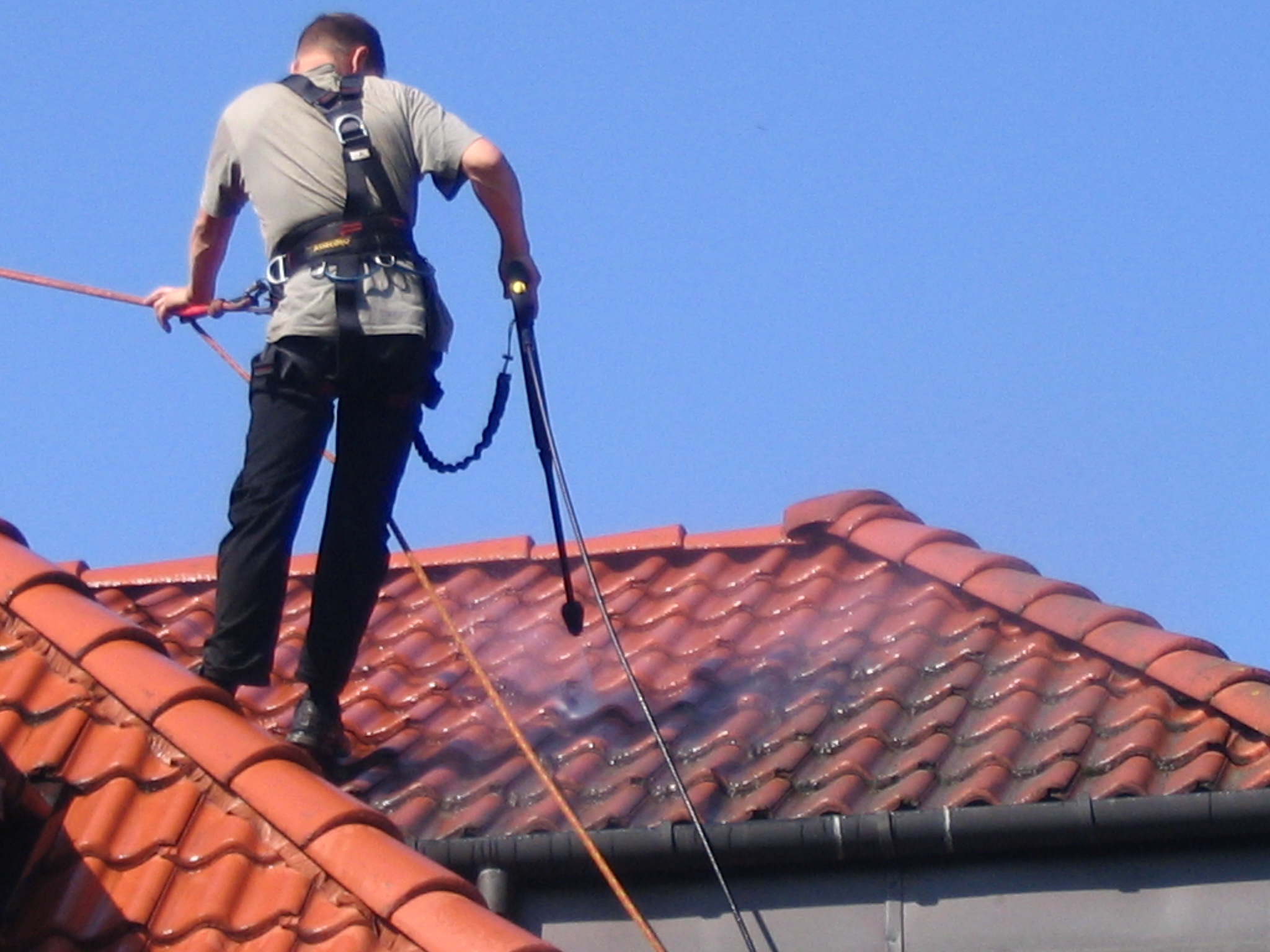Pracownik w uprzęży zabezpieczającej, myje ciśnieniowo czerwoną dachówkę, widoczny strumień wody usuwający zabrudzenia, na tle błękitnego nieba.