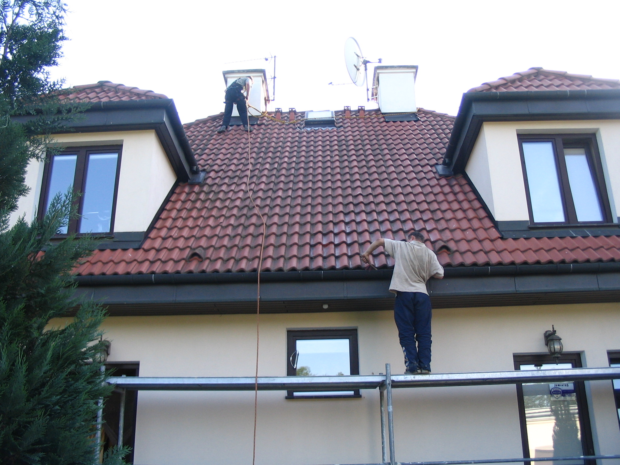 Dwóch mężczyzn na dachu domu z czerwoną dachówką, jeden na rusztowaniu przy rynnie, drugi na szczycie dachu, prawdopodobnie podczas czyszczenia lub naprawy dachu.