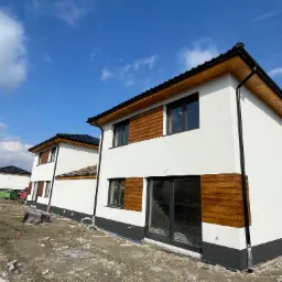 Nowoczesny dom dwukondygnacyjny z białą elewacją, drewnianymi panelami dekoracyjnymi i czarną podmurówką, widok z zewnątrz na plac budowy.