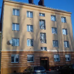 Ocieplony budynek wielorodzinny z beżową elewacją i brązowym pasem dekoracyjnym, widoczne okna z refleksami drzew, antena satelitarna i zaparkowane samochody.