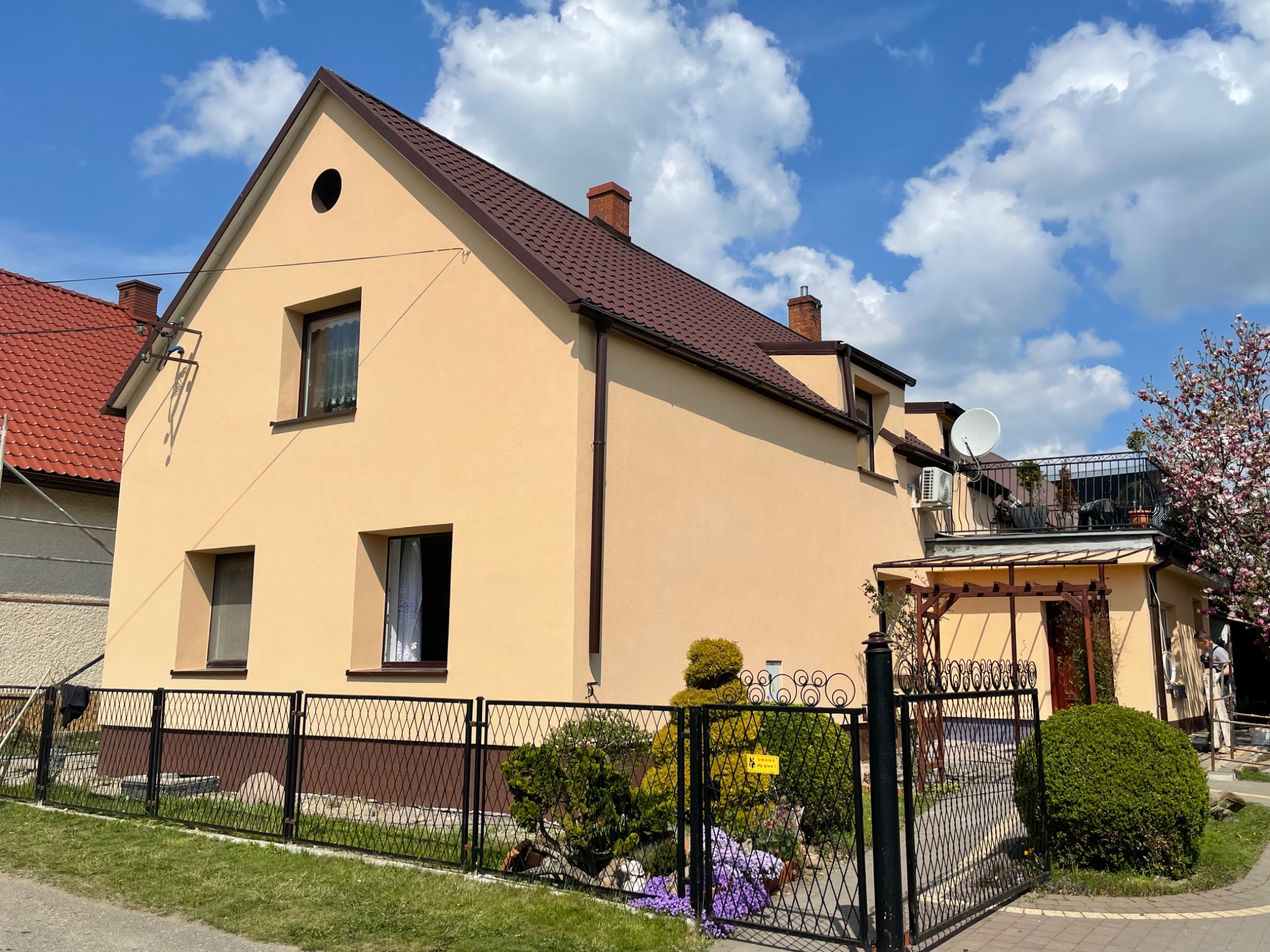 Żółty dom jednorodzinny z brązowym dachem i czarnym ogrodzeniem, widoczny z zewnątrz w słoneczny dzień, z elementami dekoracyjnymi na ogrodzeniu i zadbanym ogrodem.