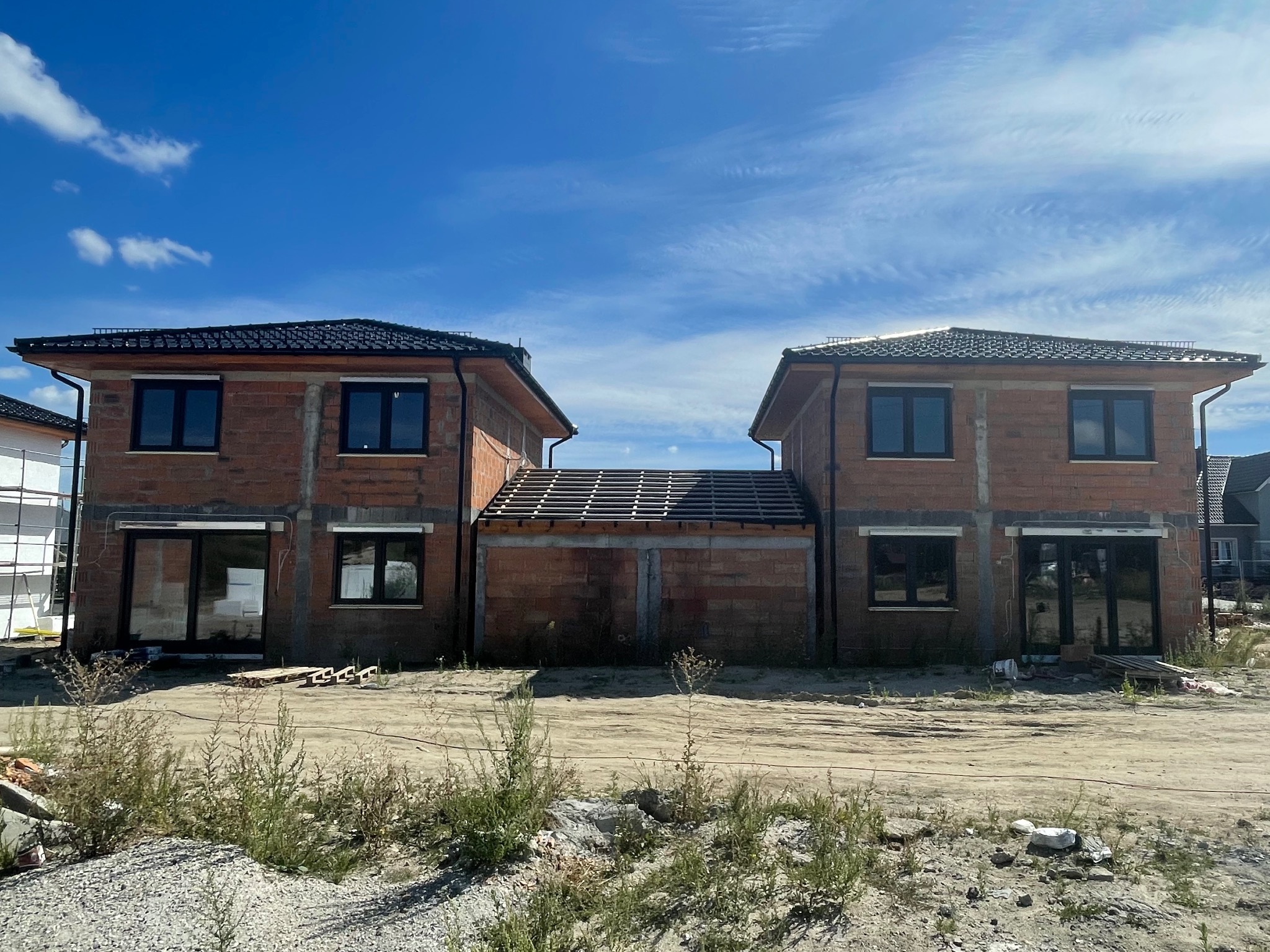 Dwa surowe, nieotynkowane domy w stanie deweloperskim połączone zadaszoną konstrukcją, z widoczną konstrukcją dachu i czarnymi oknami, na tle błękitnego nieba z chmurami.