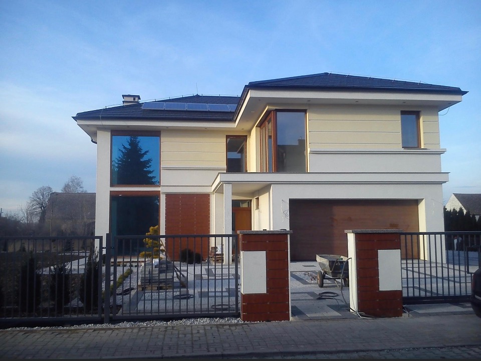 Dwukondygnacyjny dom z płaskim dachem i panelami słonecznymi, wykończony jasnym tynkiem z ciemnymi ramami okiennymi, widoczny przez metalowe ogrodzenie z elementami z drewna.