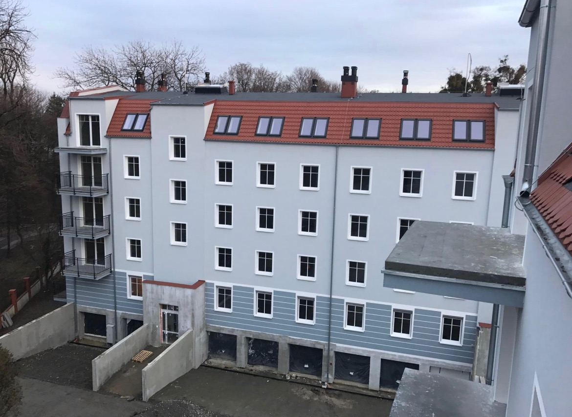 Nowo wybudowany, czteropiętrowy budynek mieszkalny z balkonami, szarą elewacją i czerwoną dachówką, widok z góry na teren wokół budynku.