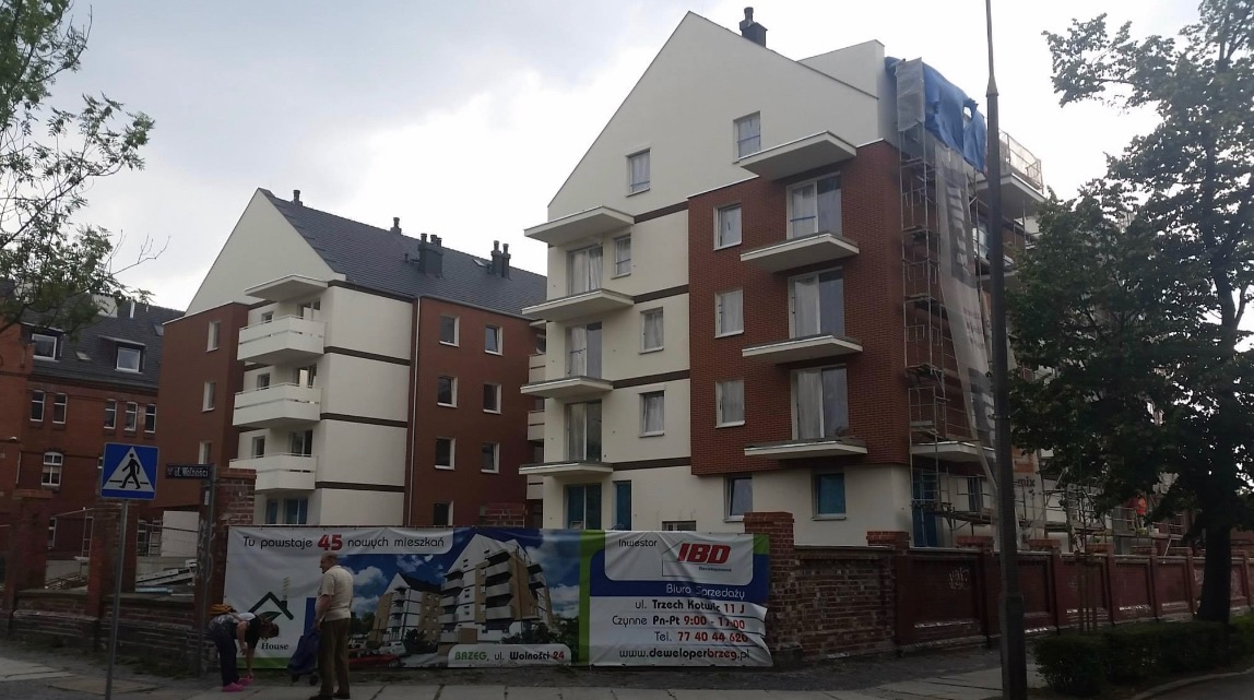 Budynek wielorodzinny z częściowo wykonaną elewacją w kolorach bieli i ceglanej czerwieni, fragment elewacji zakryty rusztowaniem i plandeką, w tle inne budynki mieszkalne.