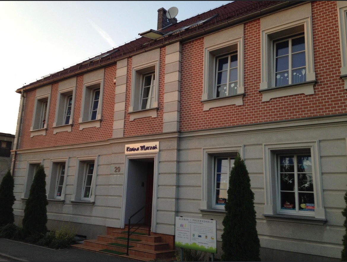 Dwupiętrowy budynek z czerwonej cegły z jasnymi obramowaniami okien i napisem 'Kraina Marzeń' nad wejściem, numer 29, widoczny szyld przedszkola.