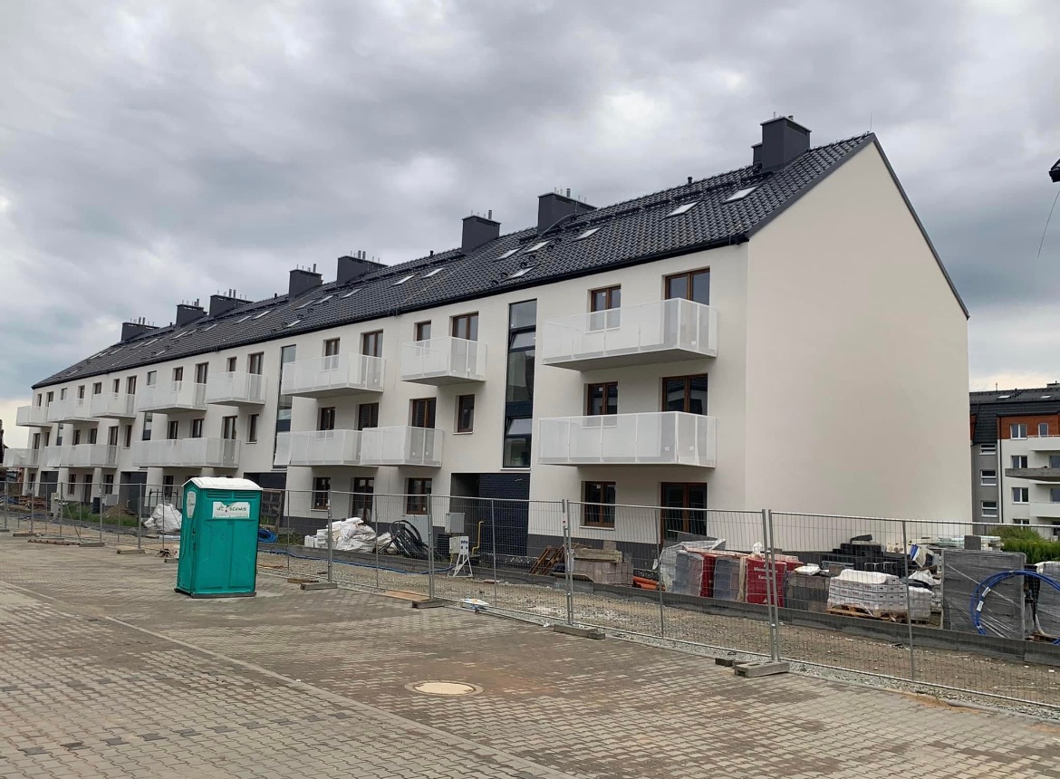 Nowoczesny budynek wielorodzinny z białymi balkonami i ciemną dachówką, widoczny na etapie wykończeniowym z tymczasową toaletą budowlaną i materiałami budowlanymi za ogrodzeniem.