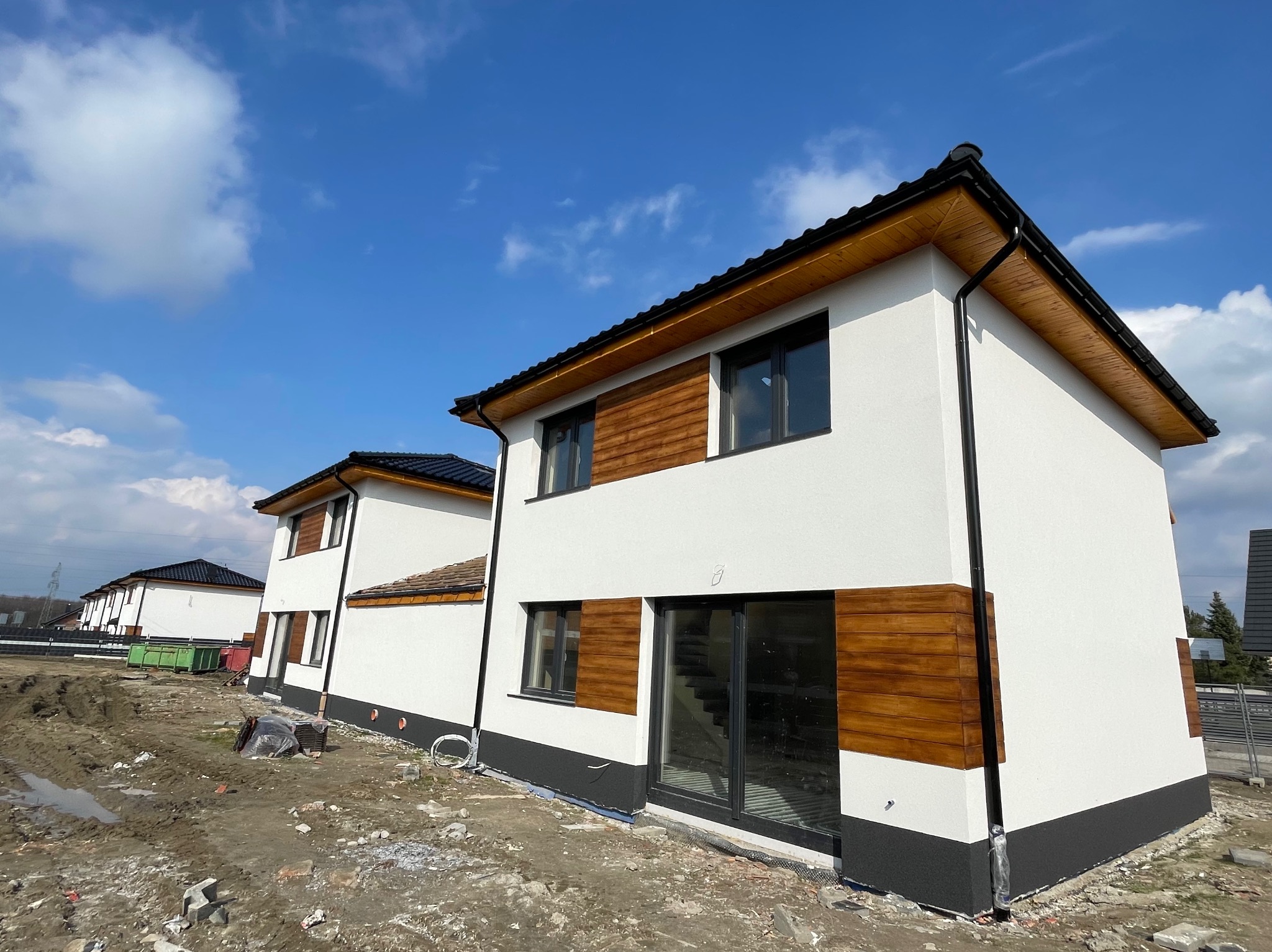 Nowoczesny dom dwukondygnacyjny z białą elewacją, drewnianymi panelami dekoracyjnymi i czarną podmurówką, widok z zewnątrz na plac budowy.