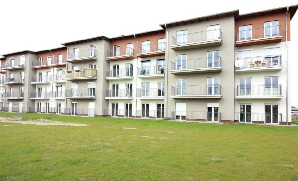 Nowoczesny budynek wielorodzinny z balkonami i zielonym trawnikiem przed fasadą.