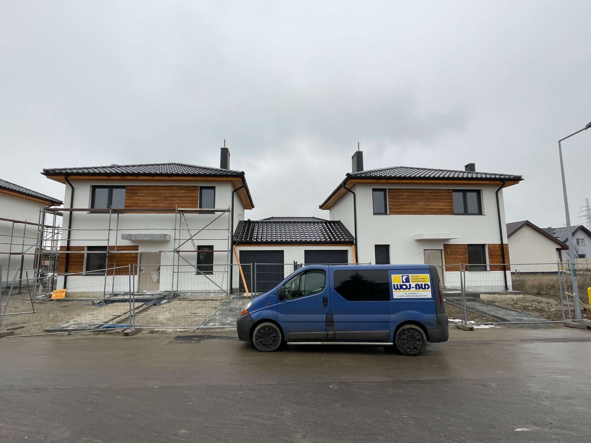 Dwa bliźniacze domy w trakcie budowy, z częściowo wykonaną elewacją i drewnianymi panelami, ogrodzone tymczasowym płotem, na pierwszym planie zaparkowany niebieski bus firmy budowlanej z widocznym...