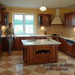 Meble kuchenne z drewna Grzegorz Jeziorski - Klasyczna kuchnia z drewnianymi szafkami, wyspą z białym blatem i widoczną osobą w tle, na podłodze wzór szachownicy.