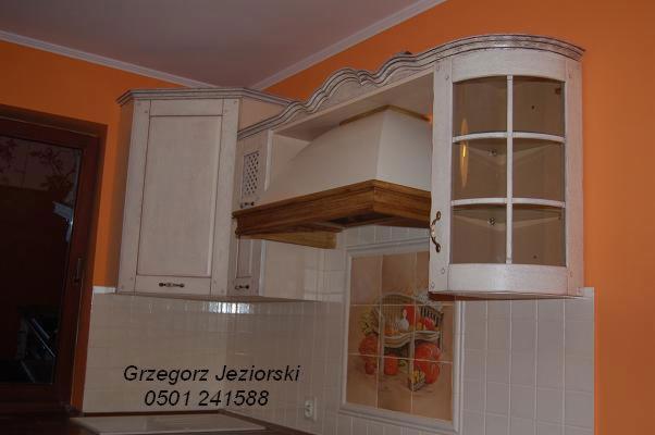 Fragment zabudowy kuchennej w stylu rustykalnym: białe szafki wiszące z dekoracyjnym frezem, okap z drewnianym wykończeniem, płytki ścienne z motywem owoców.