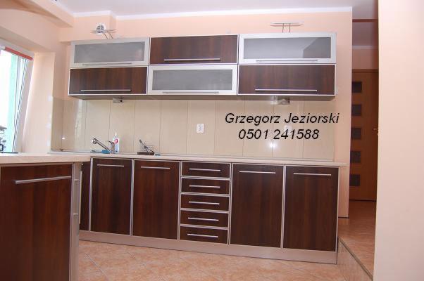 Nowoczesna zabudowa kuchenna z brązowymi frontami i aluminiowymi ramkami, górne szafki z przeszklonymi drzwiczkami, widoczny zlewozmywak i akcesoria kuchenne, jasne płytki na ścianie.