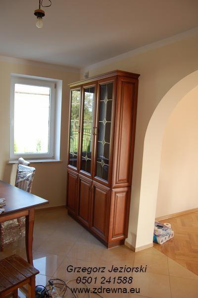 Drewniana witryna z przeszklonymi drzwiami i dekoracyjnymi szprosami, stojąca w rogu pokoju z widocznym łukiem wejściowym i częścią stołu z krzesłem w folii ochronnej.