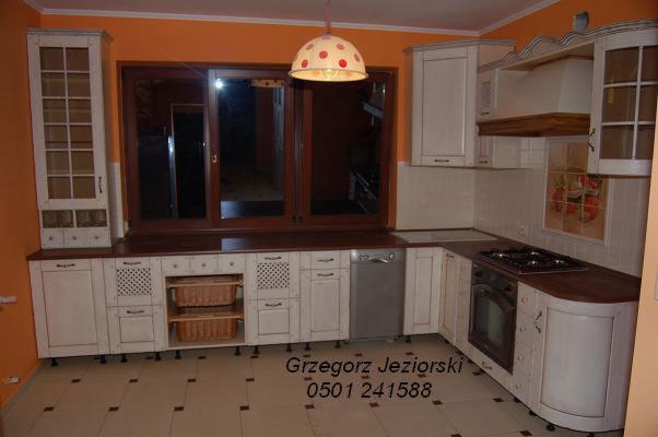 Jasna kuchnia w stylu rustykalnym z drewnianymi blatami, zabudowana pod oknem z widocznymi koszykami w szafkach i lampą w kropki.